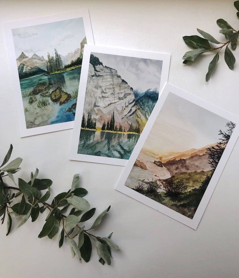May include: Three rectangular art prints featuring watercolor landscape paintings. The first shows a mountain lake with rocks, the second a steep cliff reflecting in water, and the third a golden-hued valley with dark green foliage in the foreground.