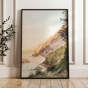 May include: A framed watercolor painting depicting a coastal landscape with a steep, grassy hillside sloping down toward a calm ocean. The scene features soft golden light, muted green and brown tones, and dark silhouettes of trees and shrubs.