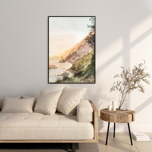 May include: A framed watercolor landscape painting depicting a coastal cliffside at sunset with soft hues of orange, brown, and green. The artwork features a rugged shoreline meeting the ocean, with dark silhouettes of trees and shrubs in the foreground.