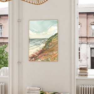 May include: A framed vertical wall art print featuring a watercolor landscape painting of a coastal scene. The artwork depicts a sandy beach meeting the ocean under a pale sky, with a grassy, textured cliffside in shades of green and brown.