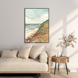 May include: A framed vertical wall art print featuring a watercolor landscape of a coastal scene with a sandy beach, dark blue ocean waves, and a grassy cliffside in shades of green and brown under a soft, pale blue and white cloudy sky.