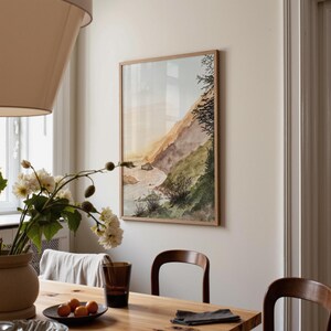 May include: A rectangular framed wall art print featuring a watercolor landscape of a coastal cliffside with green vegetation, rocky terrain, and a light sky. The artwork is displayed in a light wood frame hanging on a plain off-white wall.