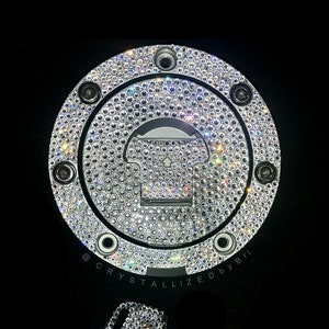 May include: A silver gas cap with a round, crystal-covered design. The cap has a small, rectangular opening in the center. The text "CRYSTALLIZED BY BRI" is visible below the cap. A silver key with a crystal-covered head is also pictured.