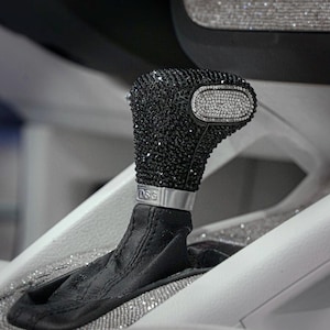 May include: Close-up of a car's interior, featuring a black gear shift knob encrusted with sparkling crystals. The shift knob has a silver band with "DSG" engraved. The surrounding console is white with crystal accents, and the dashboard has a crystal-studded trim.