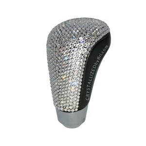 May include: A black gear shift knob covered in clear sparkling rhinestones. The text "CRYSTALLIZEDbyBri.com" is printed on the side of the knob.