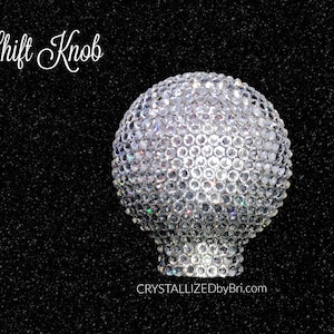 May include: A sparkling, crystal-covered shift knob. The spherical knob is covered in small, clear crystals, reflecting light. The base of the knob is also crystal-covered. The text "Shift Knob" is in white script above the knob.