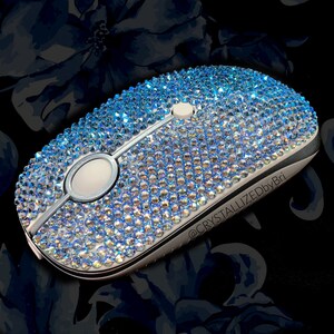 May include: A silver computer mouse covered in light blue iridescent rhinestones. The mouse is on a black background with a floral pattern. The text "@CRYSTALLIZEDbyBri" is on the side of the mouse.