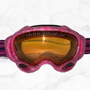 K&ouml;nnte beinhalten: Rosa Skibrille mit einem kristallbesetzten Rahmen und einer orangefarbenen, reflektierenden Linse. Die Brille hat ein schwarzes Band mit rosa und marineblauen Streifen. Der Text "CRYSTALLIZEDByBri.com" ist sichtbar.