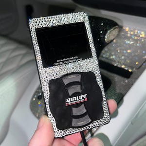 May include: A black and silver device with a screen and buttons, encrusted with rhinestones. The device has the text "AIR LIFT PERFORMANCE" and the website address "@the.diamond.eos CRYSTALLIZEDbyBri.com".