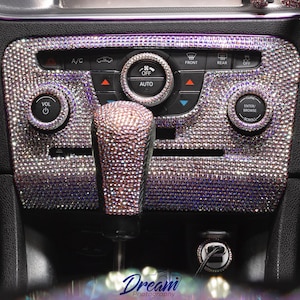 May include: Car interior detail featuring a dashboard and gear shift embellished with sparkling rhinestones. The dashboard includes controls for climate, audio, and other functions. The start button is purple with white text. The overall color scheme is silver and purple.