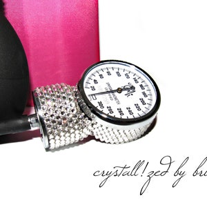 May include: A silver blood pressure gauge with a white face and black markings. The gauge is decorated with clear rhinestones. The text "SPHYGMANOMETER COIFIED 6017259" is printed on the face of the gauge. The text "crystall!zed by bri" is written in cursive below the gauge.