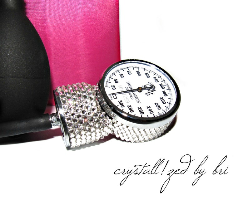 May include: A silver blood pressure gauge with a white face and black markings. The gauge is decorated with clear rhinestones. The text "SPHYGMANOMETER COIFIED 6017259" is printed on the face of the gauge. The text "crystall!zed by bri" is written in cursive below the gauge.