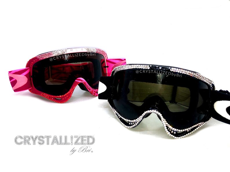 K&ouml;nnte beinhalten: Zwei Paar verzierte Skibrillen. Ein Paar ist rosa mit einem Wirbelmuster und roten Kristallakzenten. Das andere Paar ist schwarz mit silbernen Kristallakzenten. Der Text "CRYSTALLIZED by Bri" steht unten.