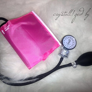 May include: A pink blood pressure cuff with a black strap and a silver gauge with rhinestones. The gauge has a black needle and a white face with black markings. The cuff has the words "Artery Mark" printed on it. The text "crystall!zed by bri" is written in black ink on a white background.