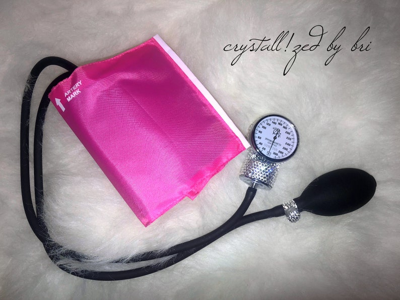 May include: A pink blood pressure cuff with a black strap and a silver gauge with rhinestones. The gauge has a black needle and a white face with black markings. The cuff has the words "Artery Mark" printed on it. The text "crystall!zed by bri" is written in black ink on a white background.