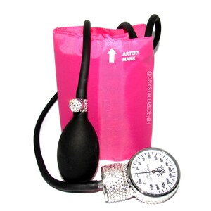 May include: A pink blood pressure cuff with a black bulb and a silver gauge covered in rhinestones. The gauge has a white face with black markings and numbers. The cuff has the text "ARTERY MARK" and "@CRYSTALLIZEDbyBri" printed on it.