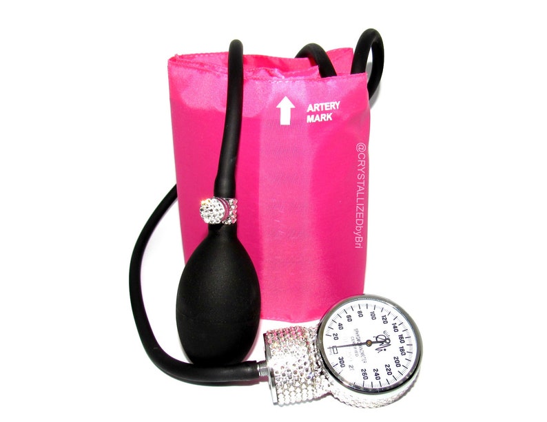 May include: A pink blood pressure cuff with a black bulb and a silver gauge covered in rhinestones. The gauge has a white face with black markings and numbers. The cuff has the text "ARTERY MARK" and "@CRYSTALLIZEDbyBri" printed on it.