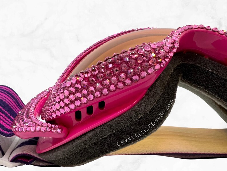 K&ouml;nnte beinhalten: Hei&szlig;rosa Brille mit rosa Strasssteinen besetzt. Die Brille hat ein schwarzes Schaumstofffutter und eine hellbraune Linse. Der Text "CrystallizedByBri.com" ist sichtbar. Ein gestreiftes Stoffband ist befestigt.