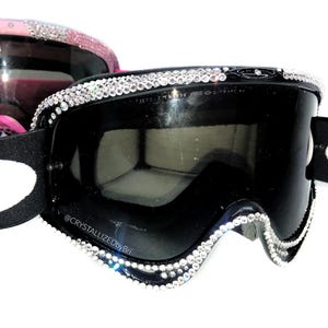 K&ouml;nnte beinhalten: Schwarze Skibrille mit kristallbesetztem Rahmen. Die Brille hat eine schwarze Linse und ein wei&szlig;-schwarzes Band. Ein zweites Paar rosa Brillen mit Kristallakzenten ist im Hintergrund zu sehen.