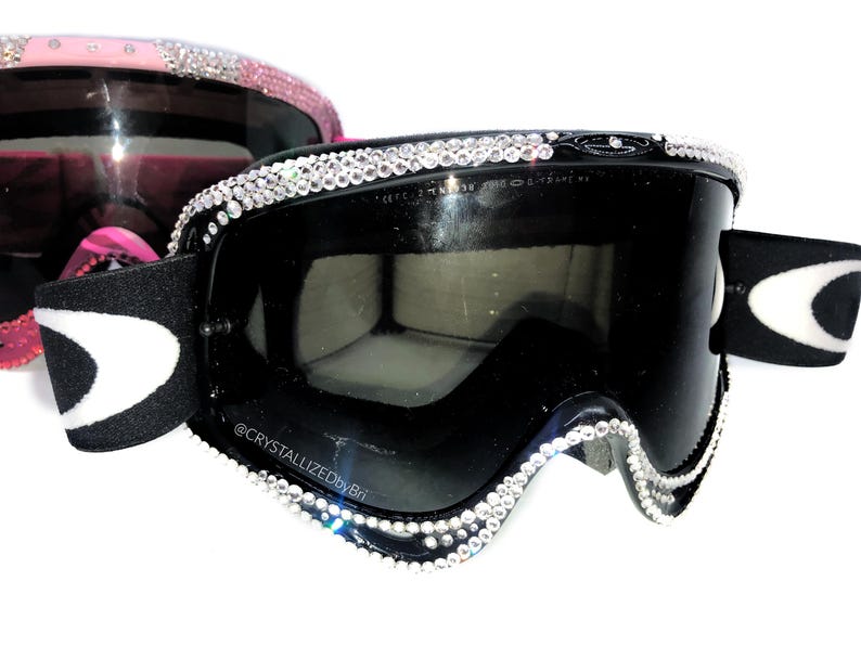 K&ouml;nnte beinhalten: Schwarze Skibrille mit kristallbesetztem Rahmen. Die Brille hat eine schwarze Linse und ein wei&szlig;-schwarzes Band. Ein zweites Paar rosa Brillen mit Kristallakzenten ist im Hintergrund zu sehen.