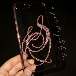 May include: Clear phone case with a rose gold frame and a pink crystal design. The design is an abstract swirl of crystals. The text "crystallized by bri" is written in cursive on the right side of the case.
