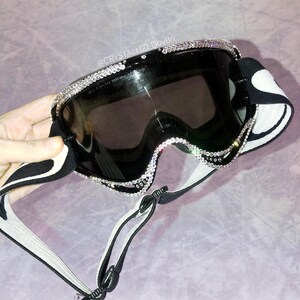 K&ouml;nnte beinhalten: Schwarze Skibrille mit dekorativem Strassbesatz um die Gl&auml;ser. Die Brille hat ein schwarz-wei&szlig; gemustertes Band und eine dunkel get&ouml;nte Linse. Der Text "@CRYSTALLIZEDbyBri" ist auf dem Bild zu sehen.