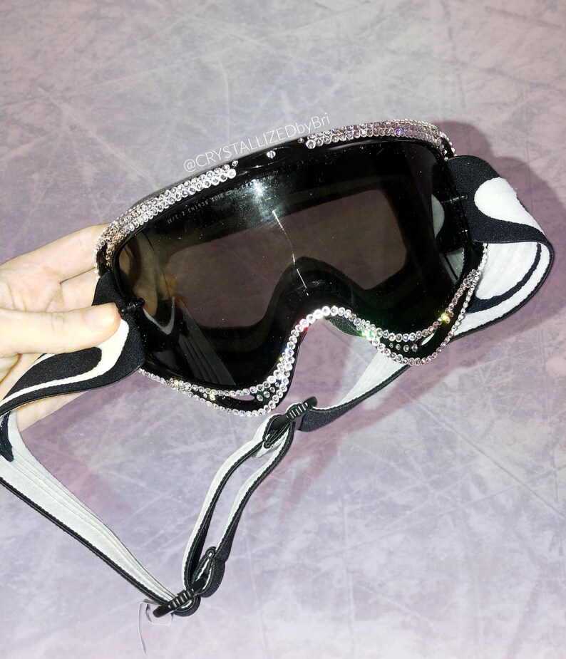 K&ouml;nnte beinhalten: Schwarze Skibrille mit dekorativem Strassbesatz um die Gl&auml;ser. Die Brille hat ein schwarz-wei&szlig; gemustertes Band und eine dunkel get&ouml;nte Linse. Der Text "@CRYSTALLIZEDbyBri" ist auf dem Bild zu sehen.