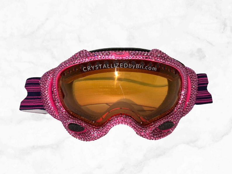 K&ouml;nnte beinhalten: Rosa Skibrille mit einem kristallbesetzten Rahmen und einer orangefarbenen, reflektierenden Linse. Die Brille hat ein schwarzes Band mit rosa und marineblauen Streifen. Der Text "CRYSTALLIZEDByBri.com" ist sichtbar.