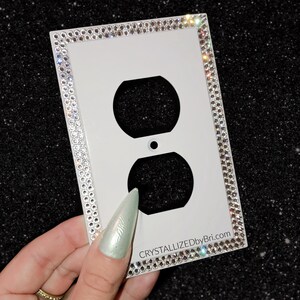 May include: White electrical outlet cover embellished with sparkling rhinestones around the edges. The cover has two circular openings and a small dot in the center. The text "CRYSTALLIZEDbyBri.com" is printed on the bottom right corner.