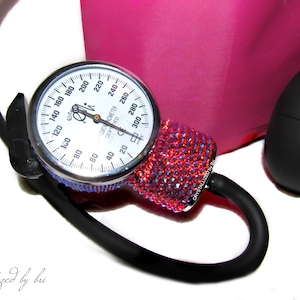 May include: A red and black blood pressure monitor with a crystal-encrusted cuff. The gauge reads up to 300 mmHg and is labeled "Sphygmomanometer Certified".
