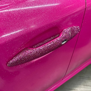 May include: A pink car door handle covered in sparkling rhinestones. The handle is labeled "CRYSTALLIZEDbyBri.com".