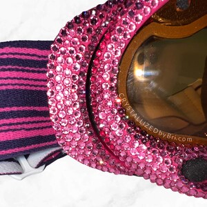K&ouml;nnte beinhalten: Nahaufnahme einer pinkfarbenen Skibrille, die mit Strasssteinen besetzt ist. Die Brille hat eine braun get&ouml;nte Linse und ein schwarz-rosa gestreiftes Band. Der Text "CRYSTALLIZEDbyBri.com" ist auf der Linse zu sehen.