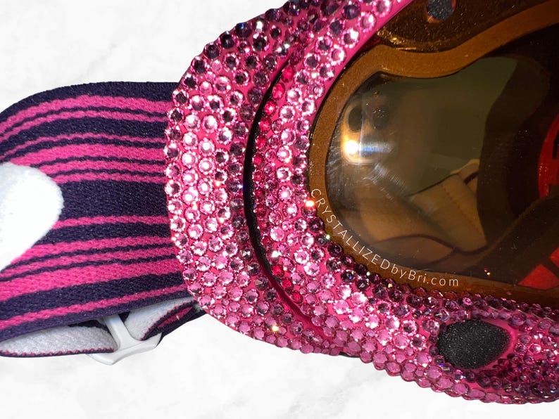 K&ouml;nnte beinhalten: Nahaufnahme einer pinkfarbenen Skibrille, die mit Strasssteinen besetzt ist. Die Brille hat eine braun get&ouml;nte Linse und ein schwarz-rosa gestreiftes Band. Der Text "CRYSTALLIZEDbyBri.com" ist auf der Linse zu sehen.