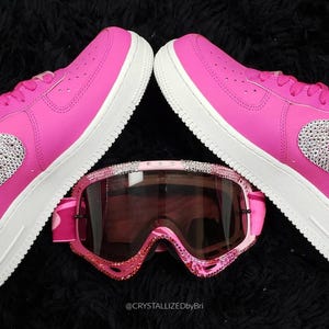 K&ouml;nnte beinhalten: Ein Paar leuchtend pinkfarbene Turnschuhe mit wei&szlig;en Sohlen und einem mit Juwelen besetzten Nike-Swoosh, kombiniert mit pinkfarbenen und mit Juwelen besetzten Skibrillen. Die Artikel sind auf einer schwarzen, strukturierten Oberfl&auml;che angeordnet. Der Text "@CRYSTALLIZEDbyBri" ist sichtbar.