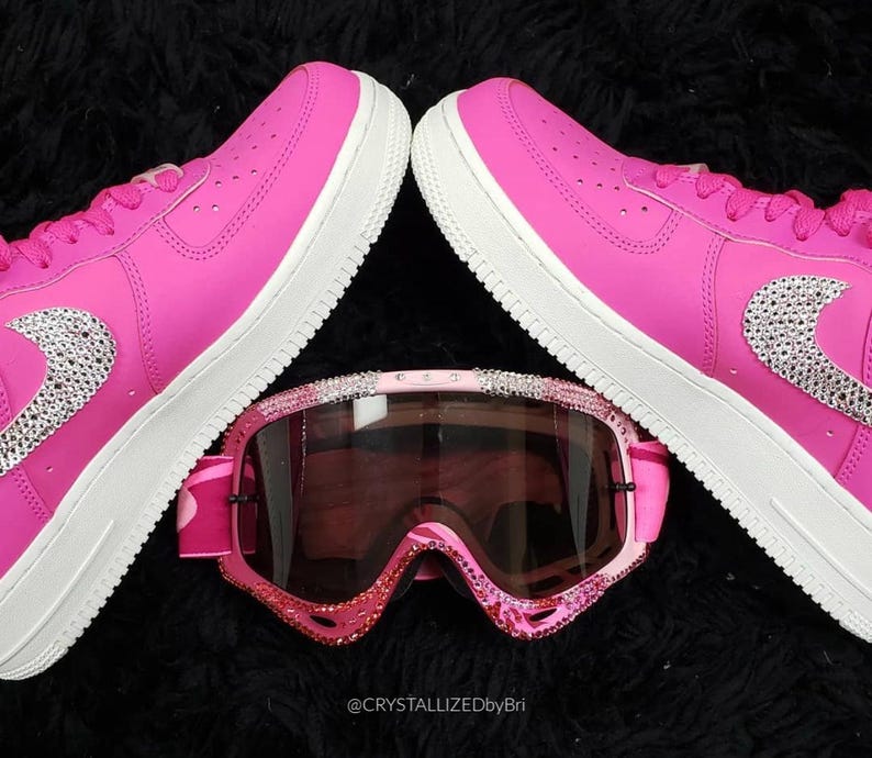 K&ouml;nnte beinhalten: Ein Paar leuchtend pinkfarbene Turnschuhe mit wei&szlig;en Sohlen und einem mit Juwelen besetzten Nike-Swoosh, kombiniert mit pinkfarbenen und mit Juwelen besetzten Skibrillen. Die Artikel sind auf einer schwarzen, strukturierten Oberfl&auml;che angeordnet. Der Text "@CRYSTALLIZEDbyBri" ist sichtbar.