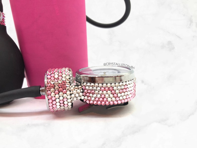 May include: A close-up of a silver medical blood pressure gauge with a pink and white rhinestone design. The gauge is attached to a black blood pressure cuff.