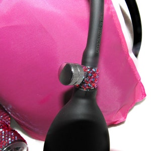 May include: A black blood pressure bulb with a silver valve and a red rhinestone band. The text "CRYSTALLIZED by B.I." is printed on the bulb. The bulb is resting on a pink fabric background.
