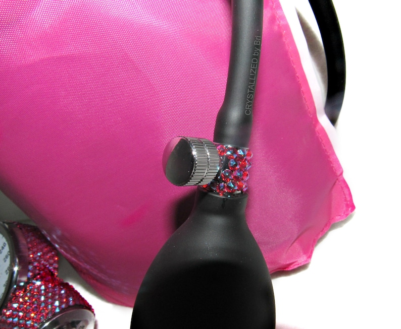 May include: A black blood pressure bulb with a silver valve and a red rhinestone band. The text "CRYSTALLIZED by B.I." is printed on the bulb. The bulb is resting on a pink fabric background.
