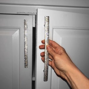 May include: Silver cabinet pulls encrusted with sparkling crystals. The rectangular pulls are mounted on a white cabinet door. The image shows a hand holding one of the pulls. The cabinet door is closed, and the other pull is visible.