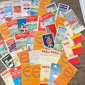 May include: A collection of vintage paper road maps from various oil companies including Shell, Esso, and Skelly, featuring colourful graphic covers with state names and regional illustrations, arranged in a fan-like pattern on a wooden surface.