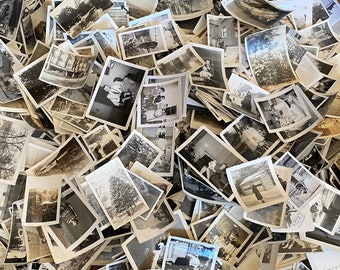 May include: A large pile of black and white photographs, some with faded edges, are stacked on a wooden surface. The photos are of various sizes and shapes, and some are slightly curled at the edges.