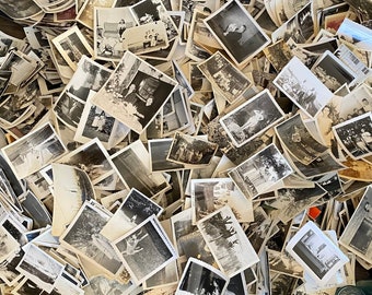 May include: A large pile of black and white photographs, some with people in them, are scattered on a wooden surface. There are also several blue glass bottles in the background.
