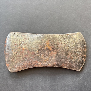 May include: A double-bit axe head made of weathered metal with a textured, pitted surface showing signs of oxidation, rust, and discoloration. The tool features two symmetrical, curved cutting edges on opposite sides of a central eye opening.