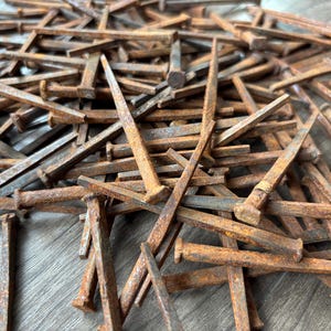 May include: A large collection of antique-style square-cut iron nails featuring a heavily rusted, textured brown finish. Each nail has a distinct flat head and a tapered, four-sided shank, scattered in a dense, overlapping pile of metal hardware.