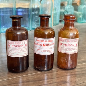 May include: Three vintage amber glass apothecary bottles with narrow necks and rounded shoulders. Each bottle features a white paper label with red text and skull-and-crossbones symbols, labeled Bichloride Mercury, Tincture of Iodides, and Benedict's Qualitative Reagent.