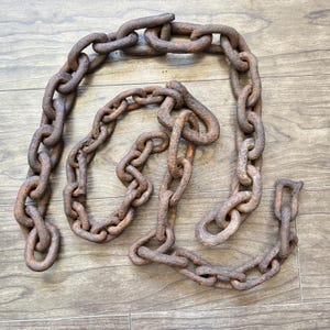 May include: A heavy, rusty metal chain with large, oval links. The chain is laid out on a wooden surface, showcasing its aged, weathered appearance. The color is a deep brown with areas of rust.