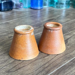 Puede incluir: Dos objetos cónicos de cerámica con un acabado marrón liso y esmaltado y una base ancha. Cada pieza presenta una abertura circular en la parte superior con un borde grueso y redondeado. Las superficies muestran variaciones sutiles en color y textura, con un interior de color claro visible en la parte superior de cada artículo, mostrando un diseño simple y funcional.