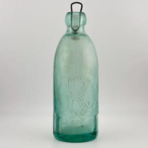 Könnte beinhalten: Eine türkisfarbene Glasflasche mit einem Metallgriff. Die Flasche hat einen geprägten Buchstaben "K" auf der Vorderseite. Sie hat einen abgerundeten Körper und einen schmalen Hals. Die Flasche ist leer und scheint antik zu sein.