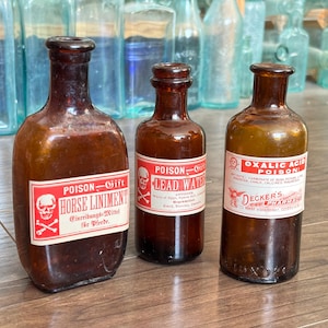 May include: Three vintage brown glass bottles with red and white labels. The labels read "POISON" and list the contents as "HORSE LINIMENT", "LEAD WATER", and "OXALIC ACID POISON". The bottles are displayed on a wooden surface.