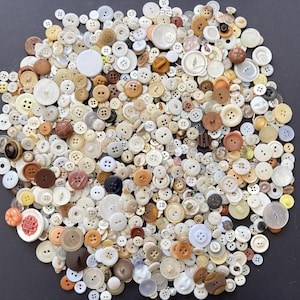 May include: A large collection of assorted sewing buttons in various sizes, shapes, and materials, including white, cream, brown, and translucent plastic, wood, and metal options, scattered across a dark surface in a dense, overlapping arrangement.